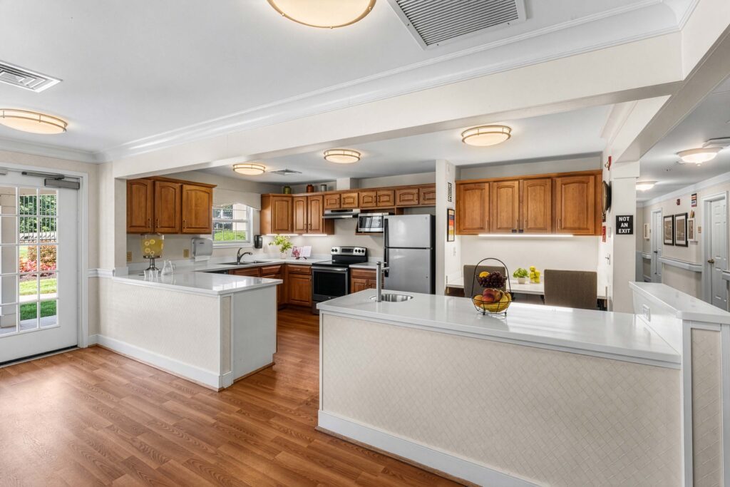 Bright communal kitchen and dining area at Artis Senior Living of Evesham memory care community, featuring wood cabinetry, a large island, and an adjoining seating nook.