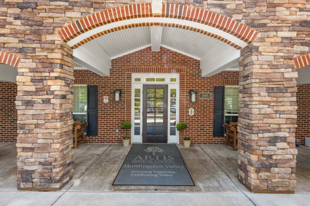 Front entrance of Artis Senior Living of Huntingdon Valley with a stone archway, brick facade, and welcome mat displaying the community's name and tagline.