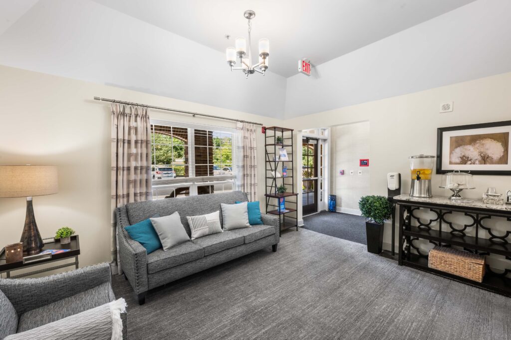 Front lobby at Artis Senior Living of Huntingdon Valley with a gray sofa, a refreshment station, and a view toward the entrance doors.