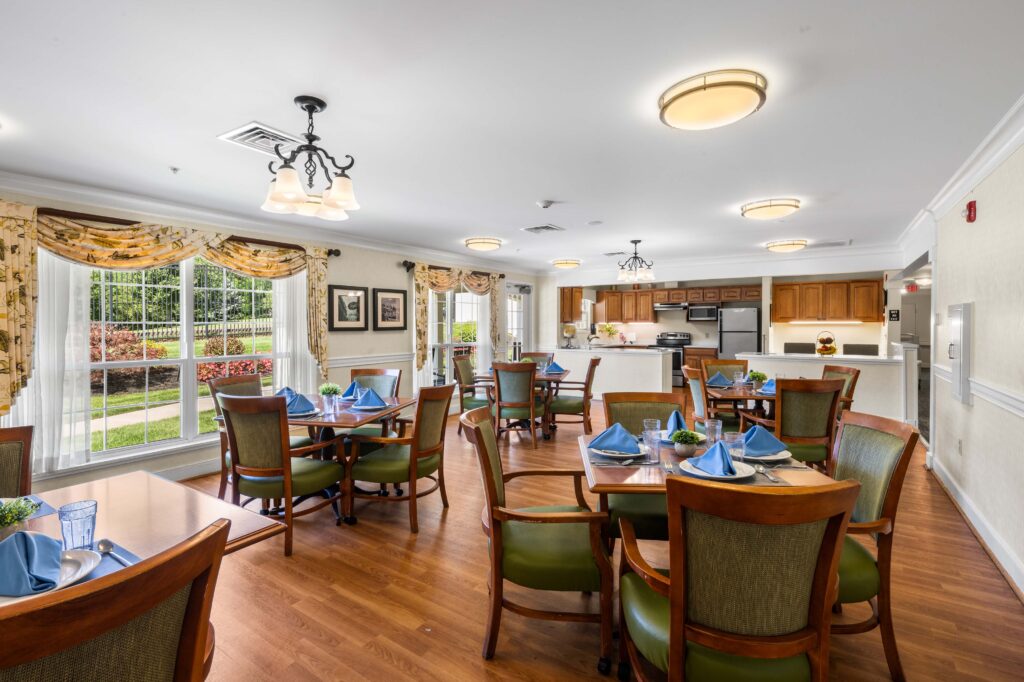 Dining room at Artis Senior Living of Huntingdon Valley with tables set with blue napkins, large windows, and an open kitchen in the background.