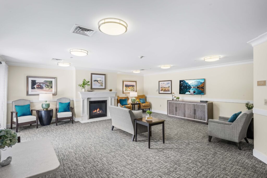 Wide view of a common living room at Artis Senior Living of Huntingdon Valley with a fireplace, TV, multiple seating areas, and framed landscape art.