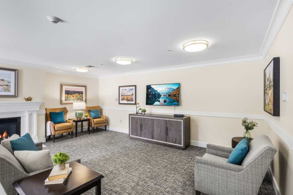 Common living room at Artis Senior Living of Huntingdon Valley with a fireplace, TV, sofas, armchairs, and framed landscape art.