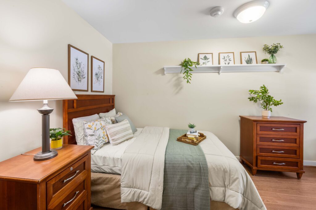 Resident bedroom at Artis Senior Living of Huntingdon Valley with a made bed, wooden headboard, nightstand lamp, dresser, and framed botanical art.