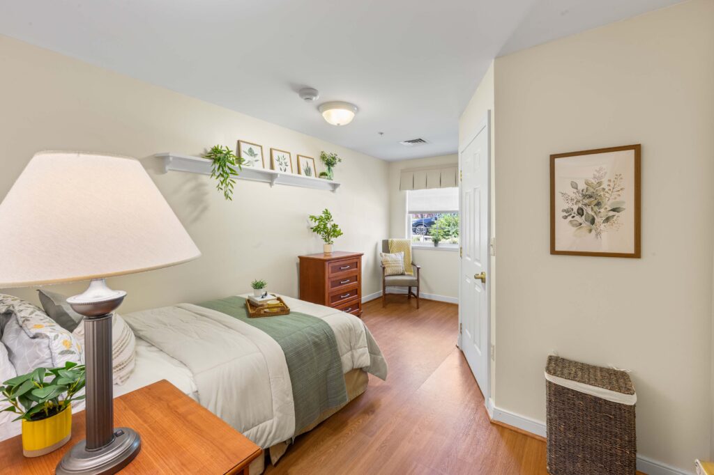Resident bedroom at Artis Senior Living of Huntingdon Valley with a made bed, nightstand lamp, dresser, armchair, and framed botanical art.