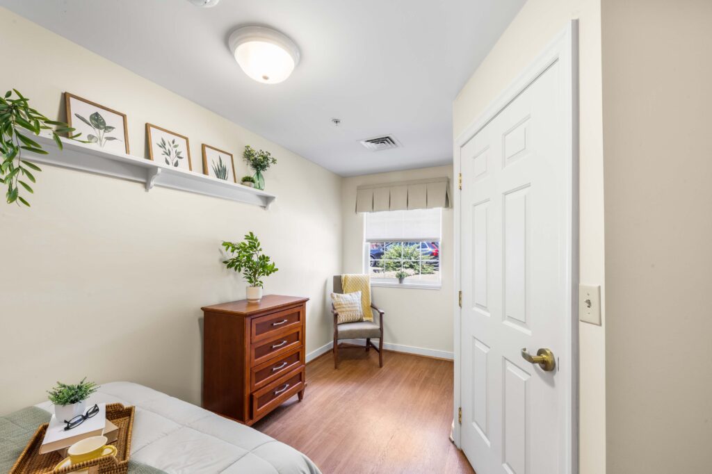 Resident bedroom at Artis Senior Living of Huntingdon Valley with a wooden dresser, armchair by a window, decorative shelf, and hardwood flooring.