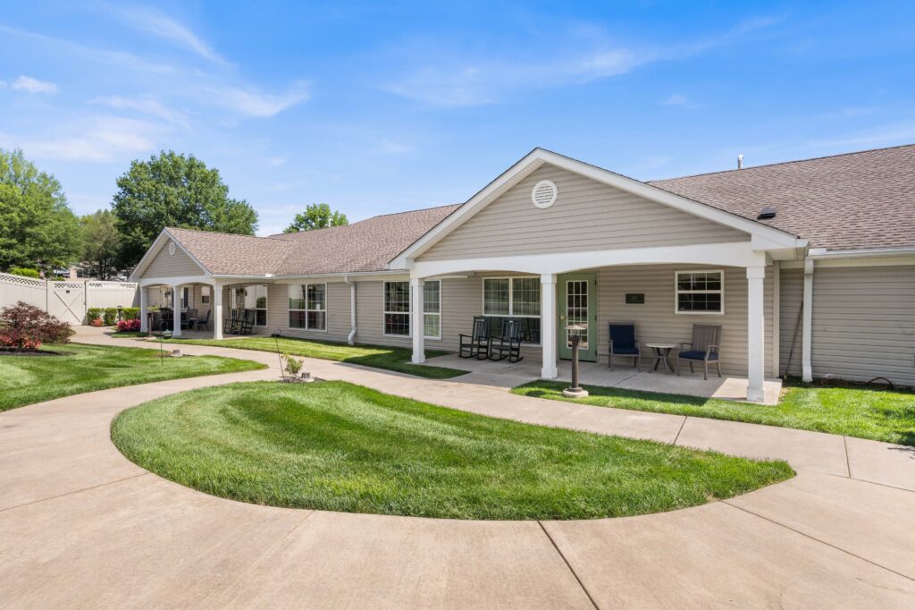 Daytime view of Artis Senior Living of Huntingdon Valley with a covered porch entrance, rocking chairs, and a curved walkway around a landscaped lawn.