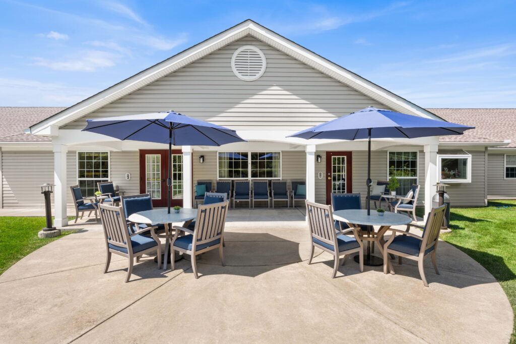 Daytime patio view of Artis Senior Living of Huntingdon Valley showing a covered patio with round tables, blue umbrellas, and lounge seating beneath the porch.