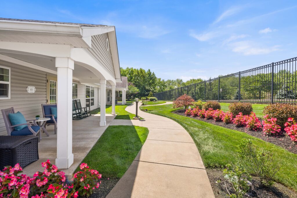 Covered porch and walking path at Artis Senior Living of Huntingdon Valley with rocking chairs, seating, and a fenced walkway lined with flowering shrubs.