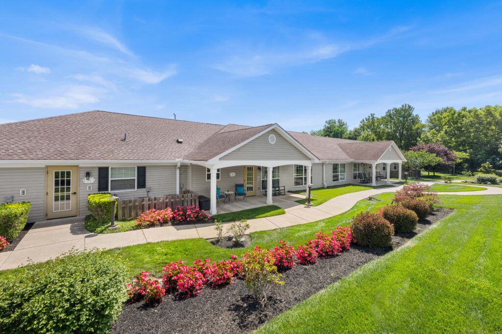 Daytime entrance of Artis Senior Living of Huntingdon Valley with a covered porch, rocking chairs, and flowering shrubs lining a curved walkway.