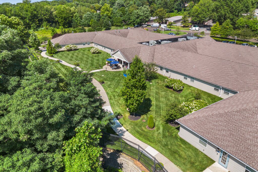 Wide aerial daytime view of Artis Senior Living of Huntingdon Valley showing the building layout, courtyard with patio seating, walking paths, and a fenced lawn surrounded by trees