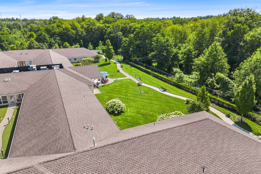Aerial daytime view of the enclosed backyard at Artis Senior Living of Huntingdon Valley with a walking path, benches, gazebo seating, and a fenced lawn bordered by trees