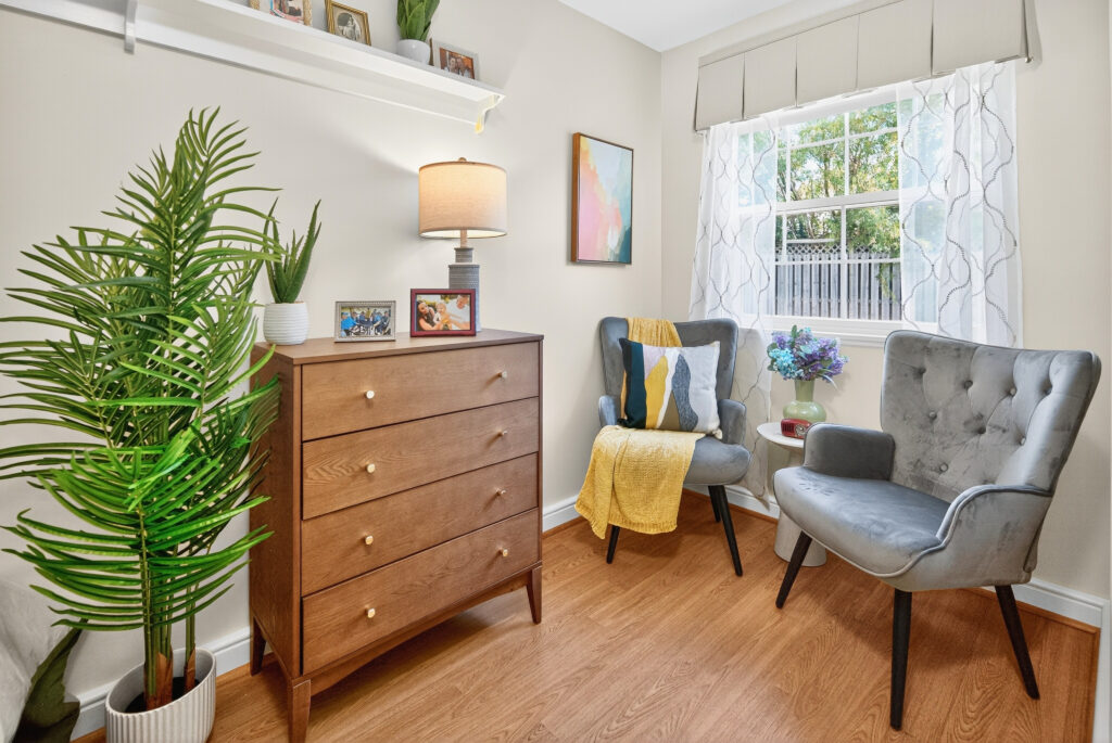 A well lit studio suite model room featuring two gray chairs accented with a yellow throw, wooden dresser, and decorative home accents