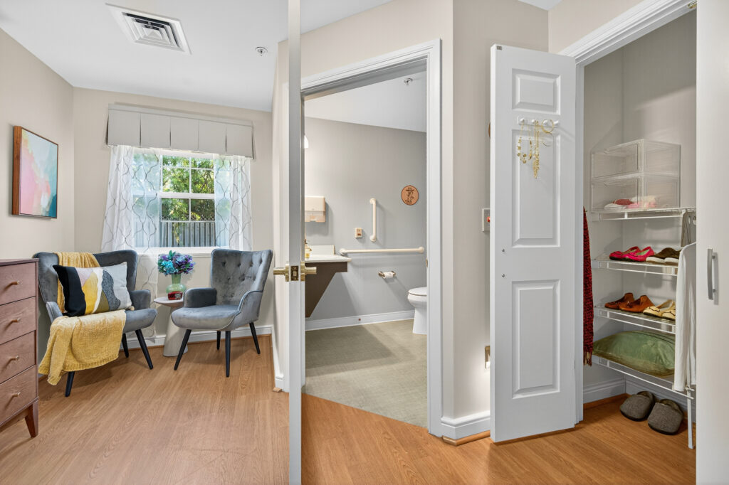 A well lit studio suite model room featuring two gray chairs accented with a yellow throw, wooden dresser, and decorative home accents