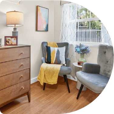A well lit studio suite model room featuring two gray chairs accented with a yellow throw, wooden dresser, and decorative home accents