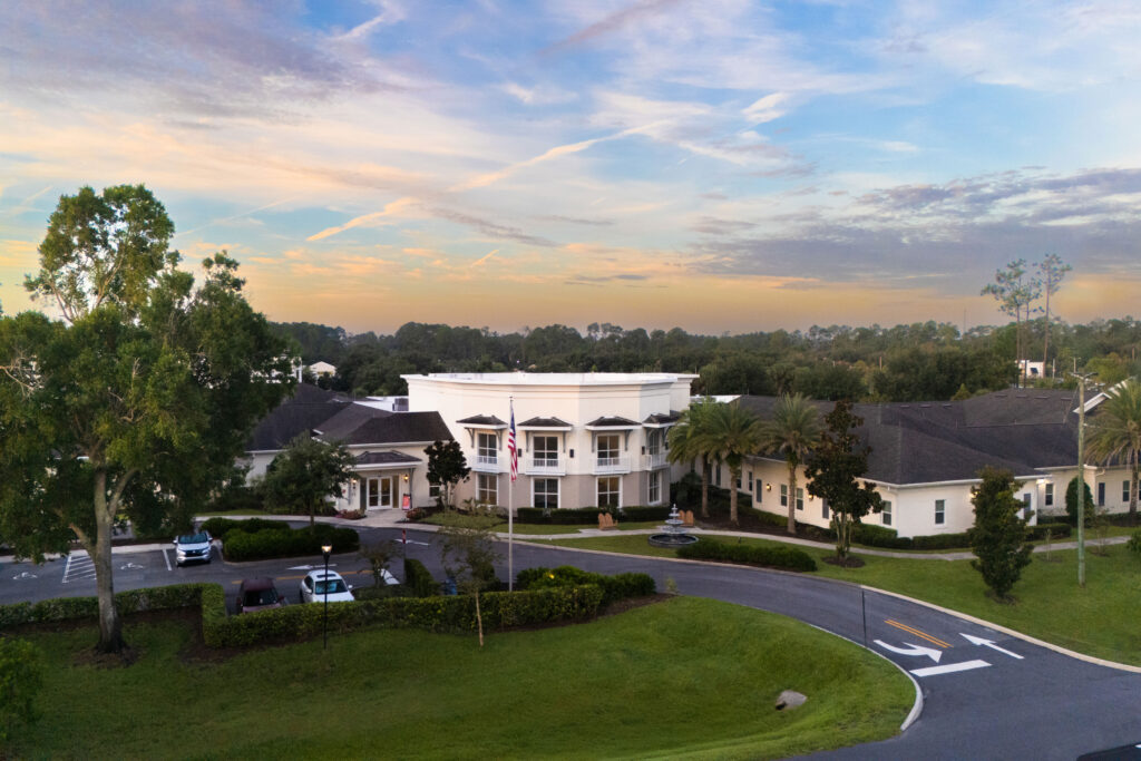 Aerial view of the Artis Senior Living of Palm Coast entrance, with circular driveway, flagpole, fountain, and monument sign along the main road