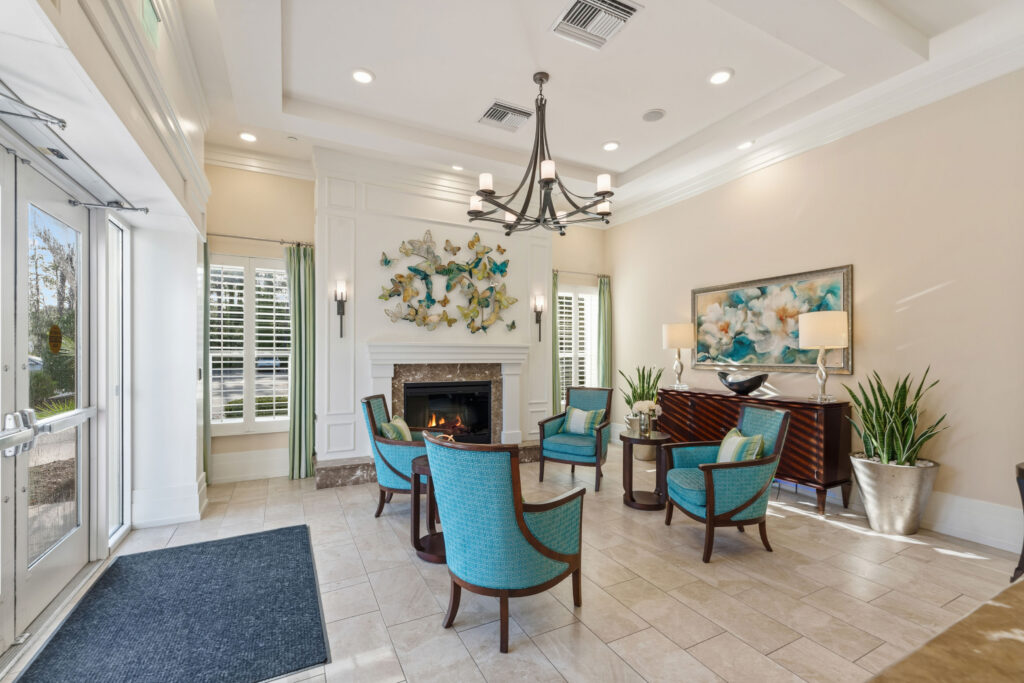 Entrance lobby at Artis Senior Living of Palm Coast with glass doors, a marble fireplace, chandelier, and teal seating area