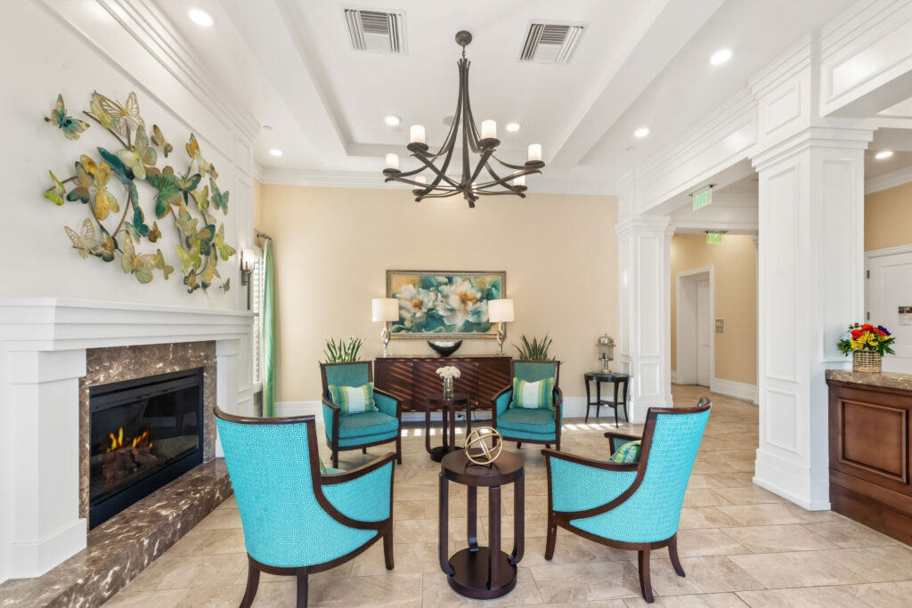 Elegant lobby lounge at Artis Senior Living of Palm Coast with a marble fireplace, chandelier, and teal armchairs arranged around a seating area