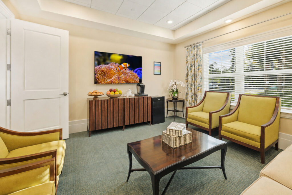Cozy lounge at Artis Senior Living of Palm Coast with a TV, coffee station, fresh fruit, and yellow armchairs