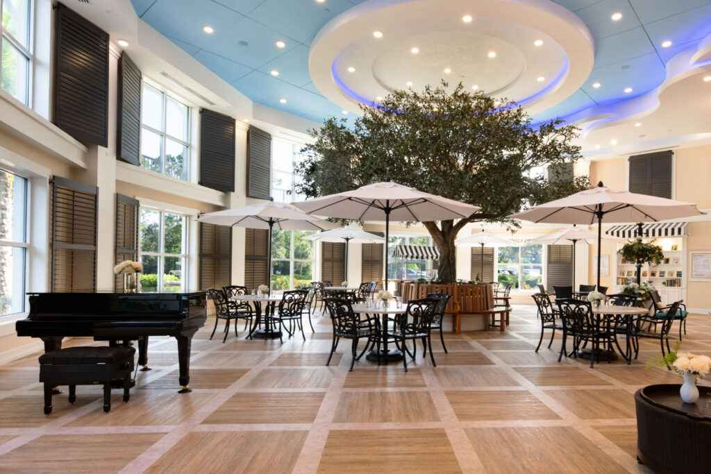 Indoor town square at Artis Senior Living of Palm Coast with a grand piano, umbrella-shaded tables, a central tree, and a sky-painted ceiling
