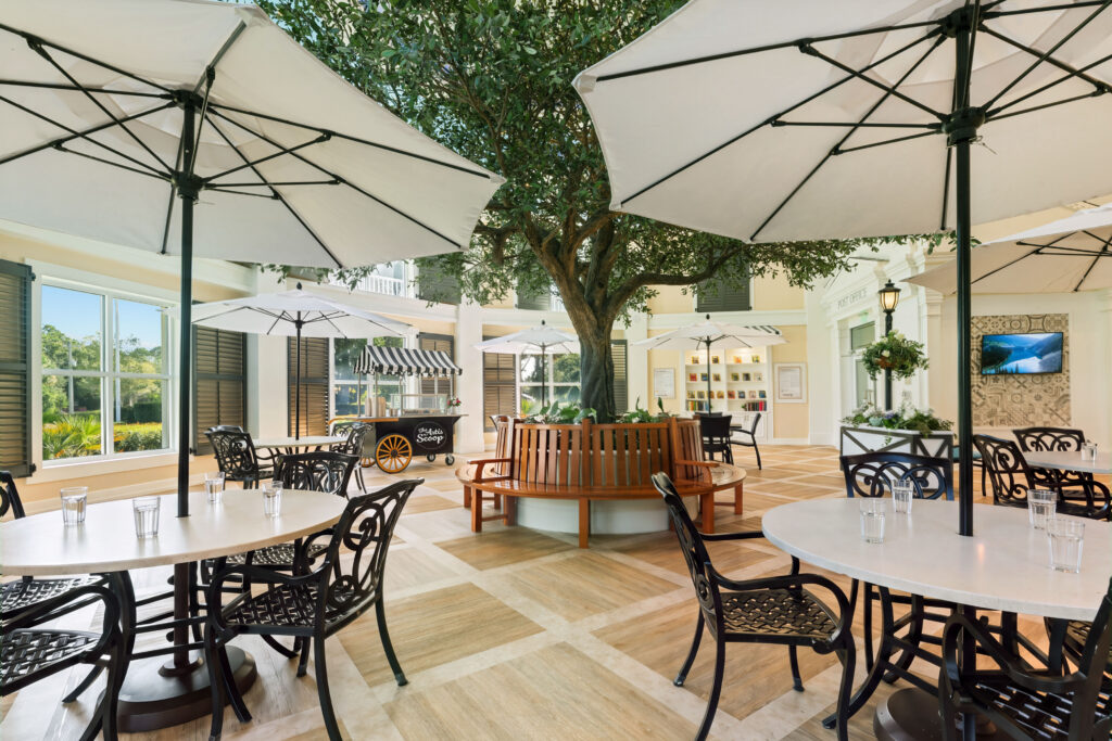 Indoor town square courtyard at Artis Senior Living of Palm Coast with a large central tree, umbrella-shaded café tables, and a post office storefront