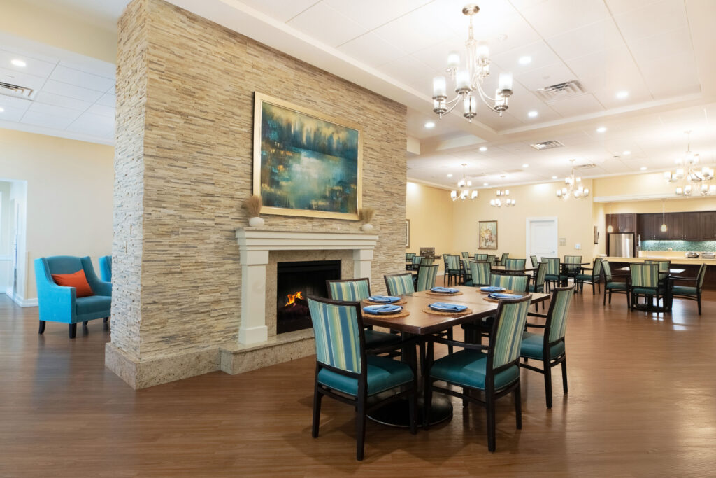 "Dining room at Artis Senior Living of Palm Coast with a set table near a stone fireplace and an open kitchen in the background