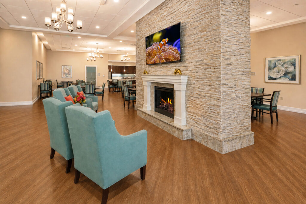 Community lounge at Artis Senior Living of Palm Coast with a stone fireplace, teal armchairs, and an open dining area in the background