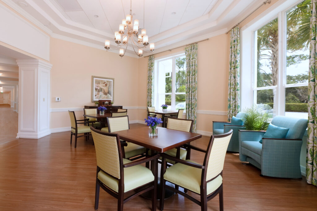 Sunroom at Artis Senior Living of Palm Coast with dining tables, a chandelier, tall windows, and blue lounge seating