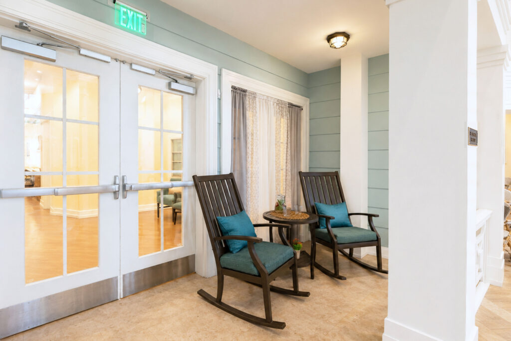 Indoor porch-style seating area at Artis Senior Living of Palm Coast with two rocking chairs and a side table near a secured exit door