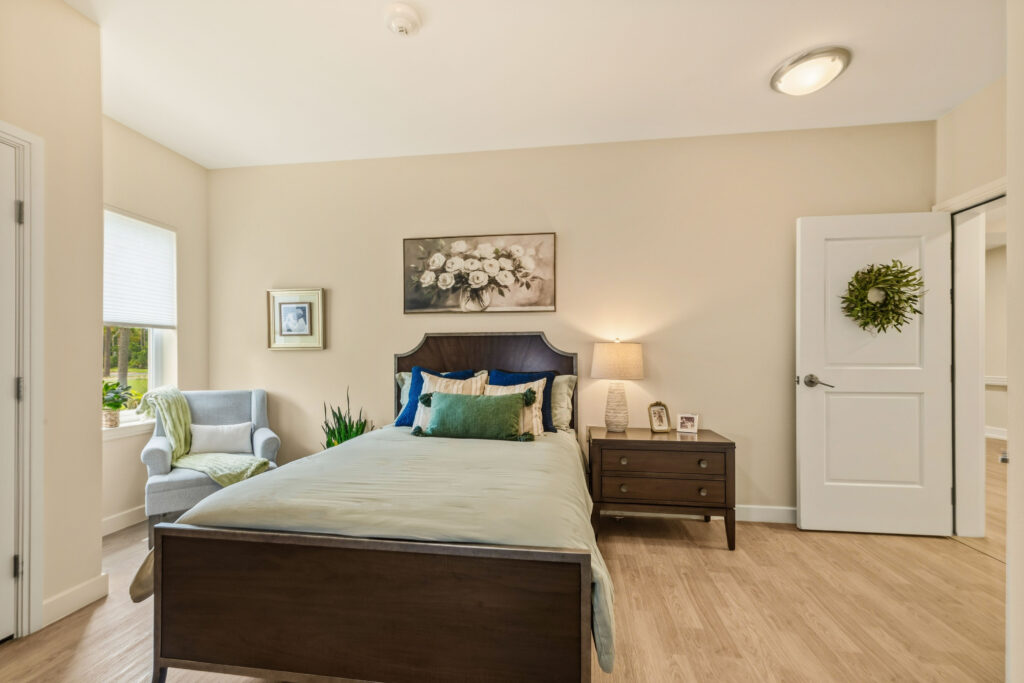 "Private memory care bedroom at Artis Senior Living of Palm Coast with a dark wood bed, nightstand, reading chair, and an open door decorated with a wreath