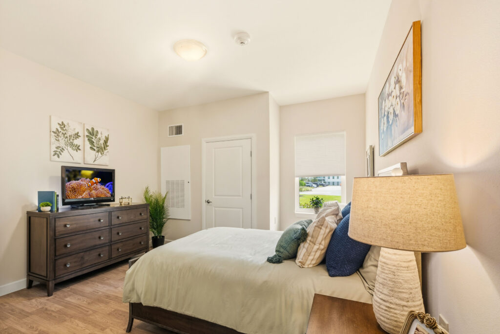 Private memory care bedroom at Artis Senior Living of Palm Coast with a queen bed, dresser with TV, and window overlooking the community grounds