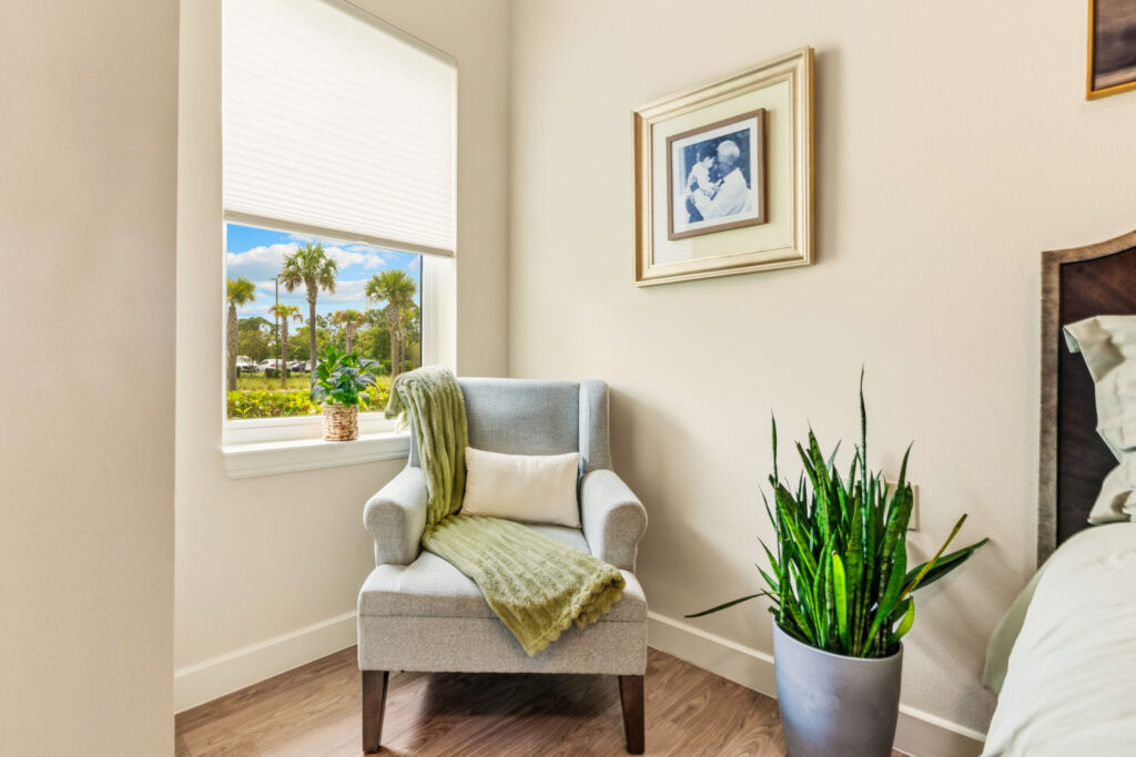 Cozy reading chair by a window in a resident bedroom at Artis Senior Living of Palm Coast, with a framed family photo and view of palm trees