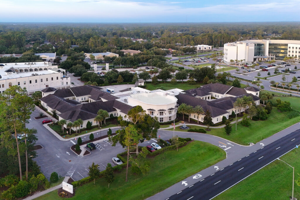 Aerial view of the Artis Senior Living of Palm Coast entrance, with circular driveway, flagpole, fountain, and monument sign along the main road