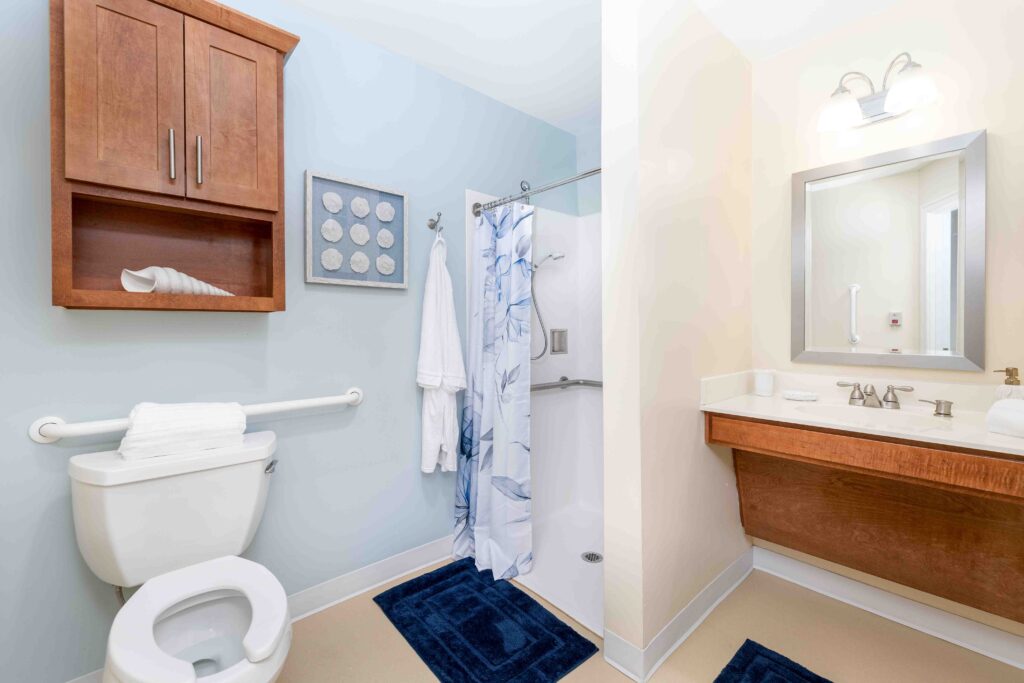 Accessible resident bathroom at Artis Senior Living of Branford with grab bars, walk-in shower, wall-mounted vanity, and coordinated blue and wood decor
