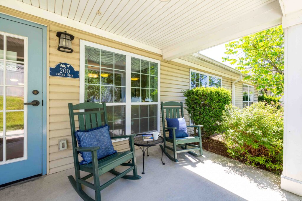 Covered porch outside the Indian Neck neighborhood entrance at Artis Senior Living of Branford, with two green rocking chairs, blue throw pillows, and a small side table