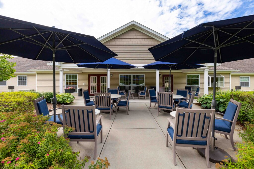 "Covered outdoor patio at Artis Senior Living of Branford with shaded umbrella tables, cushioned chairs, and landscaped shrubs leading to the community's covered porch entrance