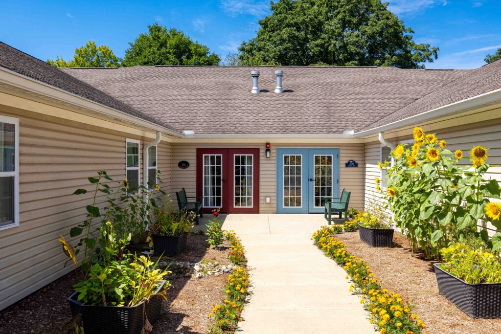 Courtyard at Artis Senior Living of Branford with red and blue French door entrances, sunflowers, marigolds, and a paved walking path between neighborhoods