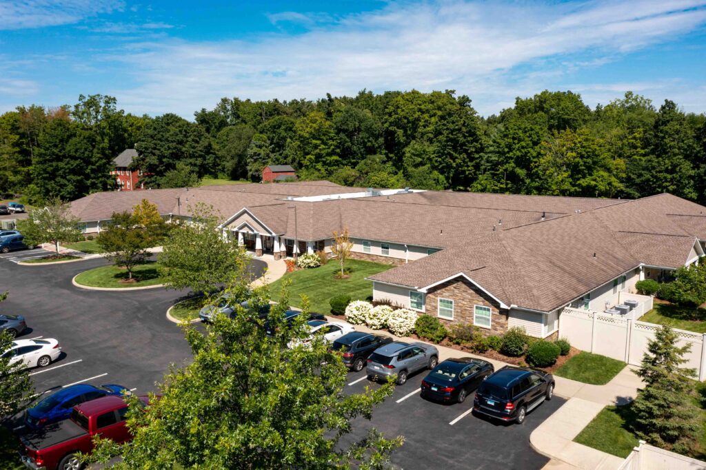 Aerial view of Artis Senior Living of Branford memory care community showing side entrance, resident parking lot, and stone-accented single-story building bordered by trees