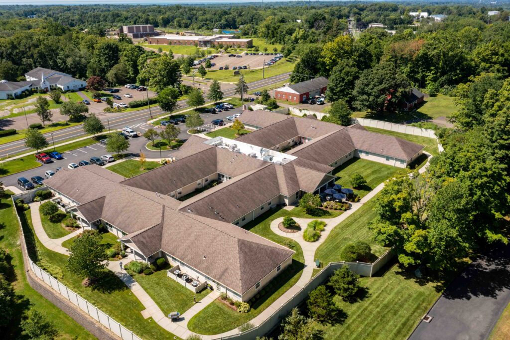 Aerial view of Artis Senior Living of Branford memory care community showing single-story building layout, secure fenced courtyard, walking paths, and outdoor patio surrounded by trees