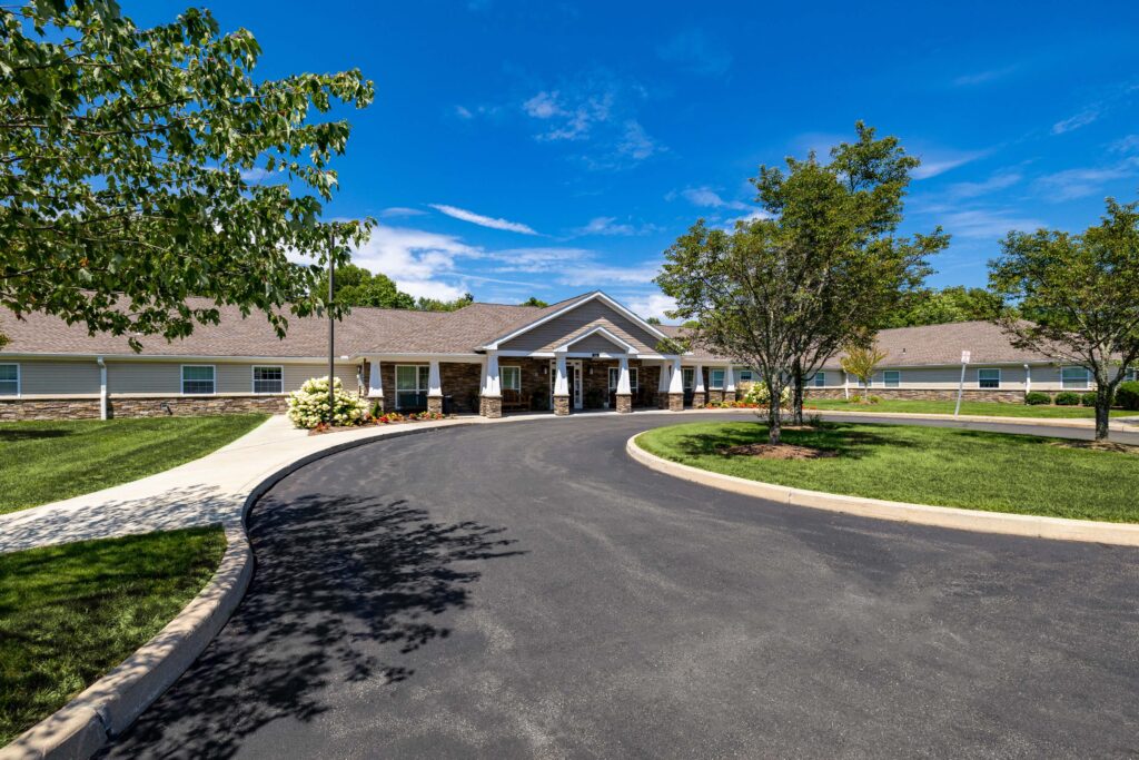 Daytime front exterior of Artis Senior Living of Branford with a curved driveway, manicured lawn, flowering hydrangeas, and a stone-and-siding craftsman-style building