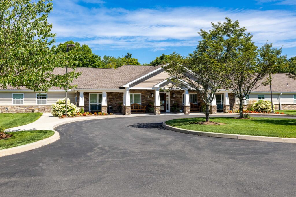 Daytime front exterior of Artis Senior Living of Branford with a curved driveway, manicured lawn, flowering hydrangeas, and a stone-and-siding craftsman-style building