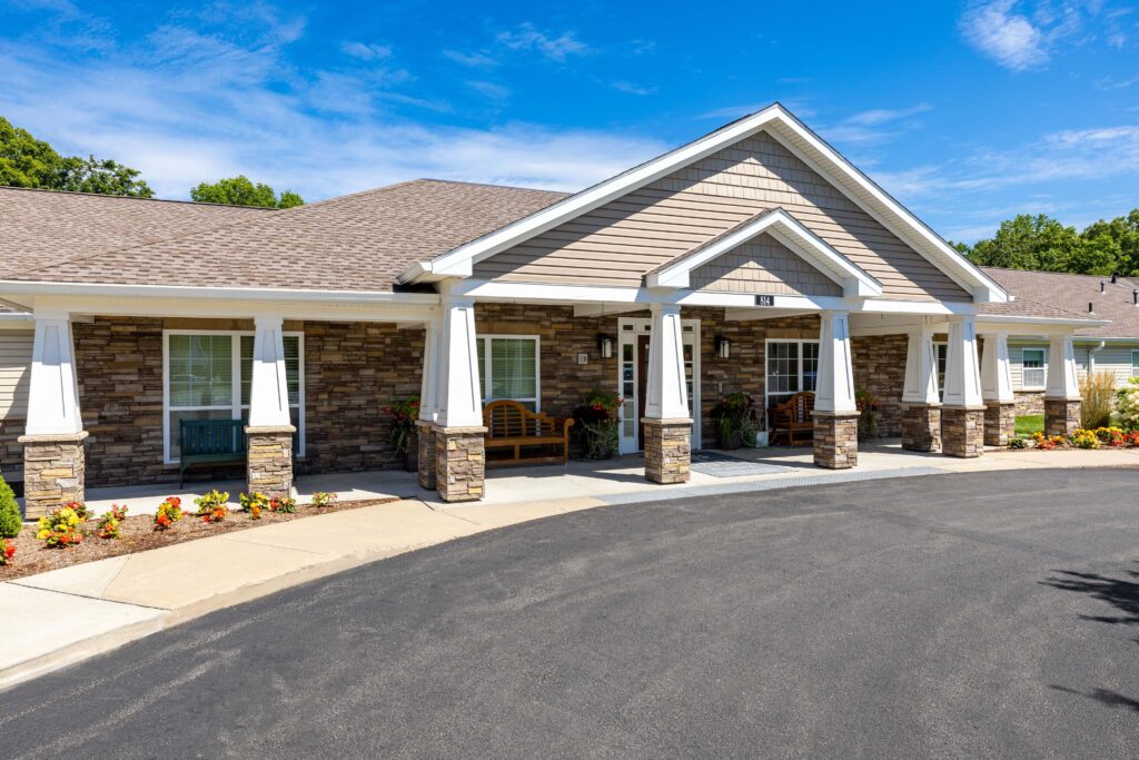 Wide view of the front entrance at Artis Senior Living of Branford, 814, showing an extended covered porch with stone columns, wooden benches, and seasonal flower beds along the walkway