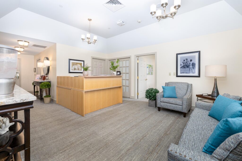 Reception lobby at Artis Senior Living of Branford with a wood front desk, community map, teal and gray sofa seating, chandelier, and vintage black-and-white photography