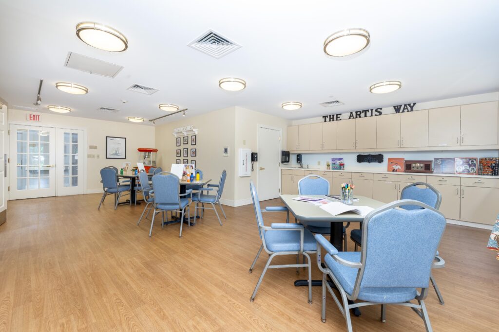 Activities room at Artis Senior Living of Branford with 'The Artis Way' signage above cabinetry, a demonstration kitchenette, vintage radio, and group tables with blue chairs