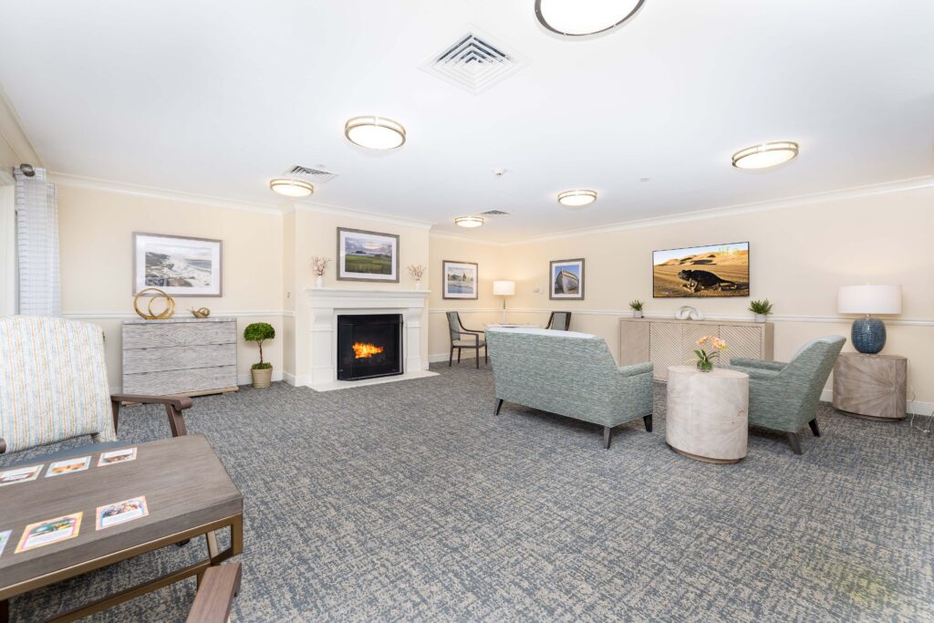 Combined living room and TV lounge at Artis Senior Living of Branford with a lit fireplace, sage green seating, wall-mounted TV, and a card table with playing cards in the foreground