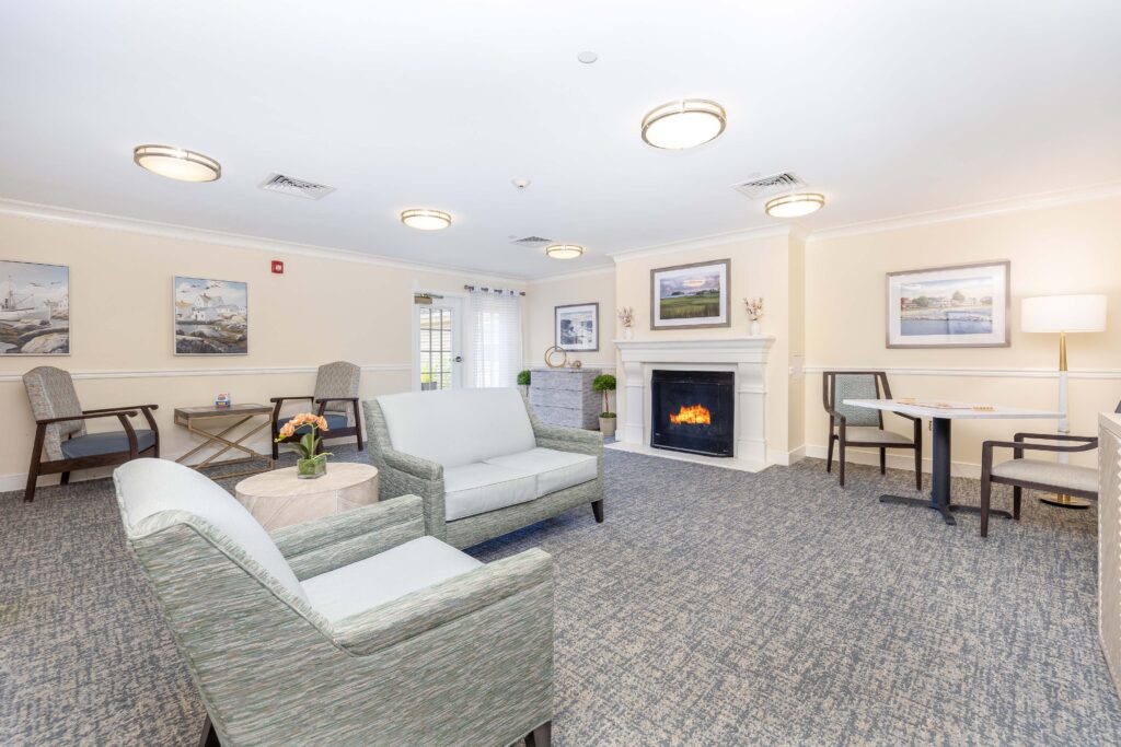 Living room at Artis Senior Living of Branford with a lit fireplace, sage green sofa and armchairs, coastal artwork, and a small game table for residents to gather