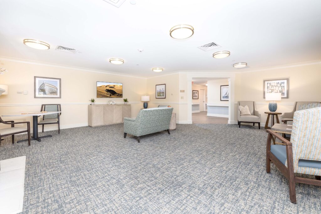 Open common area at Artis Senior Living of Branford with multiple seating groups, wall-mounted TV, bistro table, and coastal artwork, opening into a connecting hallway