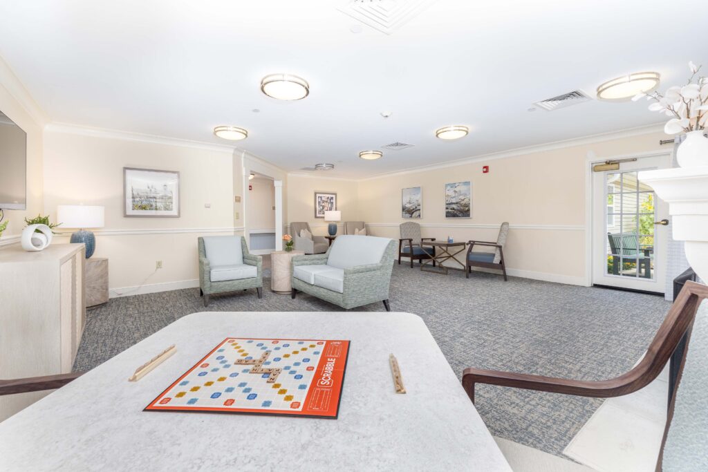 Activity lounge at Artis Senior Living of Branford with a Scrabble board set up on a game table, soft blue-green seating, and a door leading to an outdoor patio
