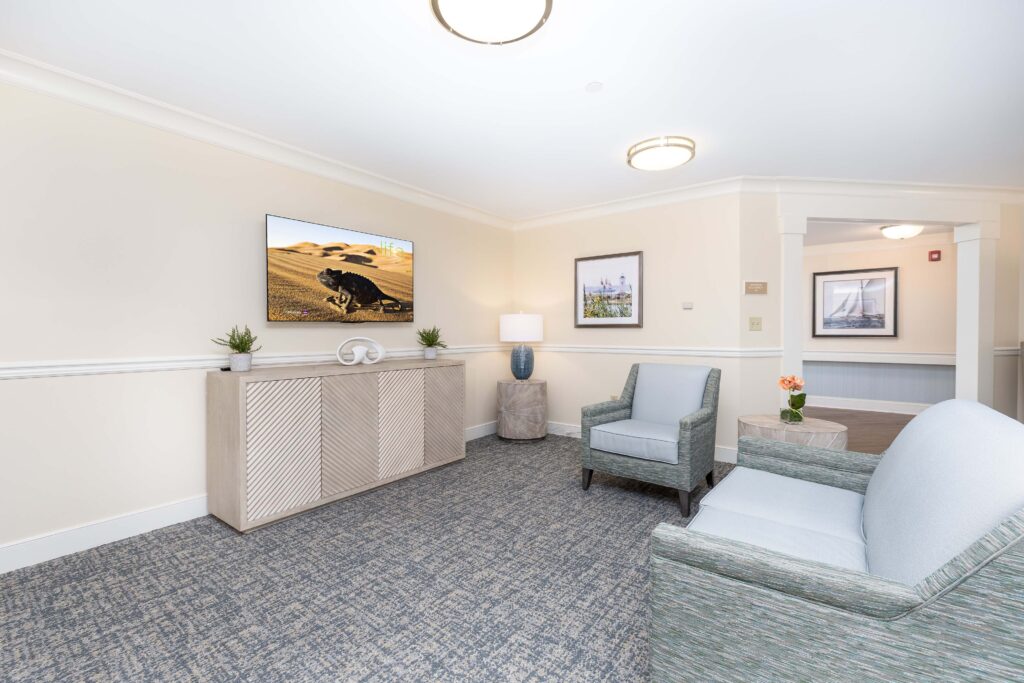 Common area lounge at Artis Senior Living of Branford with a wall-mounted TV, wooden media console, blue upholstered armchairs, and a sofa arranged for group seating