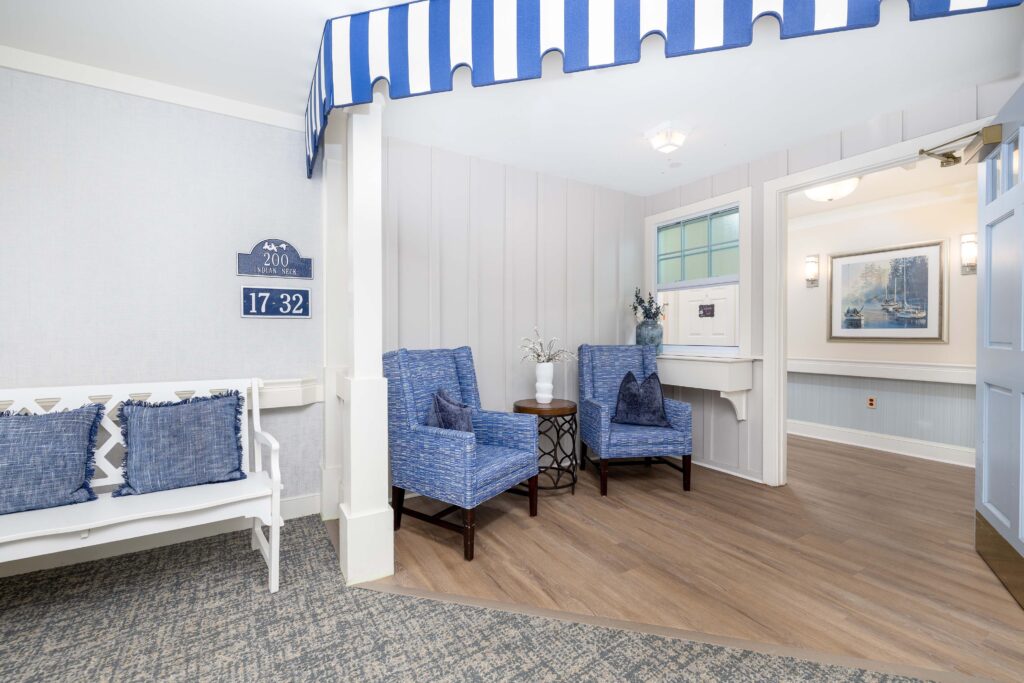 Interior sitting nook inside the Indian Neck neighborhood at Artis Senior Living of Branford, with blue armchairs, a white bench, striped awning, and coastal wall art visible through the entryway