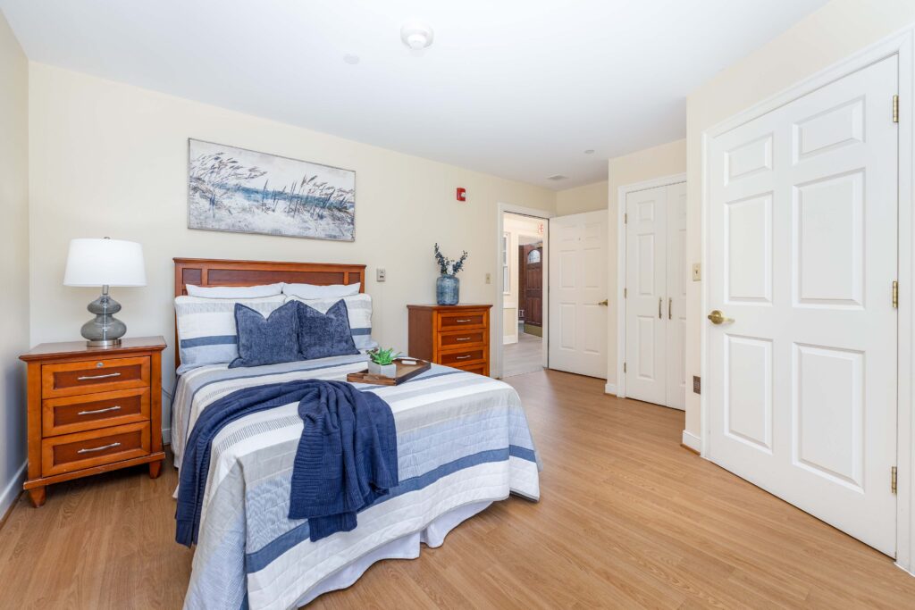 Resident bedroom at Artis Senior Living of Branford with wood bed frame, blue striped bedding, matching nightstand and dresser, double closet, and view into hallway with exit sign